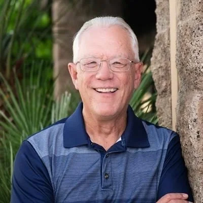 Russ Sullivan, smiling, with white hair and glasses, wearing a navy polo shirt and standing beside a stone wall with desert palms behind him
